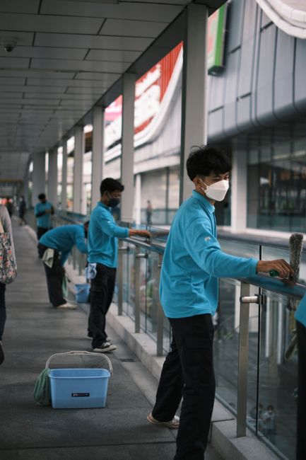 Multiple individuals wearing face masks and blue uniforms are engaged in surface cleaning activities along a glass and metal railing in a spacious, modern indoor station area. They are wiping down the
