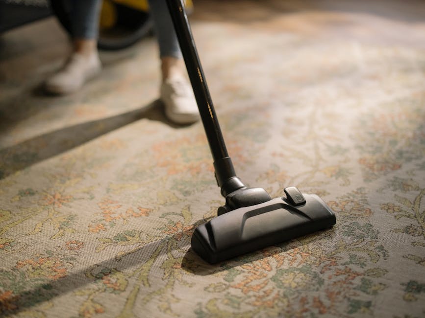 A professional cleaner dressed in white protective suit, mask, and blue gloves is using a steam cleaning machine to deep clean a beige carpet in a bright, modern living room. The room features a woode